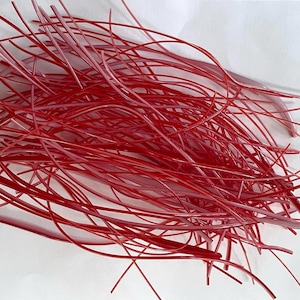 May include: A collection of thin, flexible, translucent red plastic strands scattered in a random, overlapping arrangement. The strands vary slightly in shade from deep crimson to lighter pinkish-red tones, creating a dense, tangled web-like appearance.