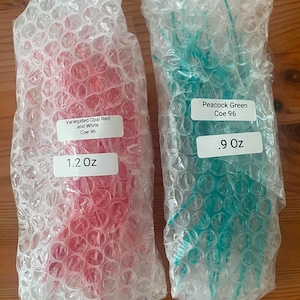 May include: Two packages of glass rods wrapped in clear bubble wrap. The left package contains variegated opal red and white glass rods weighing 1.2 oz. The right package contains peacock green glass rods labeled Coe 96 weighing .9 oz.
