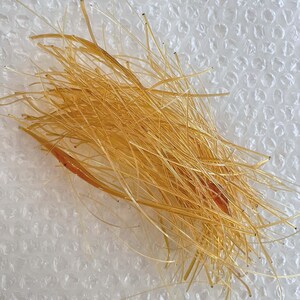May include: A close-up shot of a pile of golden-yellow, thin, thread-like material. The strands are densely packed and appear to be slightly translucent. Some strands have dark tips. A few strands have a reddish-brown hue.