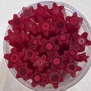May include: A collection of red, star-shaped hard candies packed vertically inside a clear, round container. Each candy features a layered, concentric design in varying shades of red, with a solid circular center surrounded by star-shaped edges.