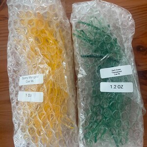 May include: Two packages of glass stringers wrapped in clear bubble wrap. The left package contains yellow glass labeled Sunny Marigold Coe 96, 1 oz. The right package contains dark green glass labeled Dark Green Transparent Coe 96, 1.2 oz.