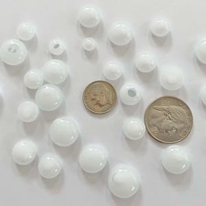 May include: White glass beads of varying sizes are scattered on a white surface, alongside two coins. The beads range from small to large, with some having holes. The coins are a dime and a quarter.