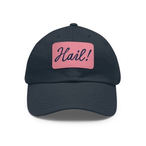 Puede incluir: Una gorra de béisbol azul marino con un parche rosa que dice "Hail!" en una fuente cursiva azul marino.