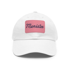 Puede incluir: Gorra de béisbol blanca con un parche rectangular rosa. El parche muestra la palabra "Manistee" en cursiva. La gorra tiene una visera curva y un diseño clásico.