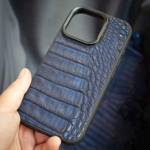 May include: A rectangular smartphone case featuring a deep blue textured surface with a pattern resembling reptile scales. The case has a solid black frame and a raised black border surrounding the square camera cutout on the upper back section.