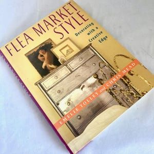 May include: A book titled "Flea Market Style" with a cream-colored cover and a purple spine. The cover features an illustration of a chest of drawers, a framed picture, and the text "Decorating with a Creative Edge."