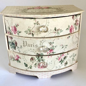May include: A three-drawer chest featuring a cream-colored base with a floral pattern of pink roses and green leaves. The middle drawer displays the word Paris in a dark font. Each drawer is equipped with a clear, round, faceted glass knob.