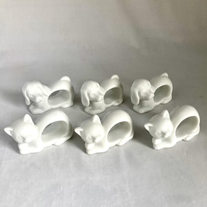 May include: Set of six white ceramic napkin rings. Three are shaped like dogs, and three are shaped like cats. Each animal has a circular opening for a napkin. The animals are in a resting position.