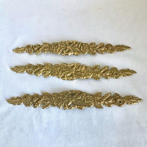 May include: Three gold-colored, ornate floral appliques. Each applique features a central arrangement of roses and leaves, with detailed textures and a symmetrical design. The appliques are ideal for decorative projects.