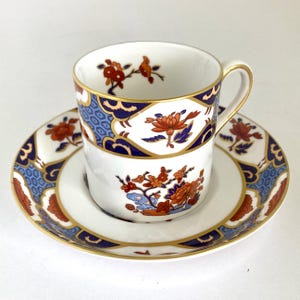 May include: A white porcelain teacup and saucer set featuring a repeating pattern of orange flowers, dark blue geometric shapes, and gold trim. The cup has a curved handle and the saucer has a wide rim decorated with the same floral and blue motifs.