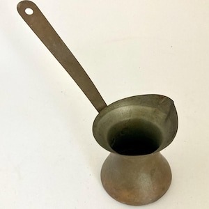 May include: A metallic coffee pot with a wide, flared opening and a long, flat handle featuring a circular hole at the end. The vessel has a rounded base and a tapered neck, displaying a weathered, brownish-grey patina across its entire surface.