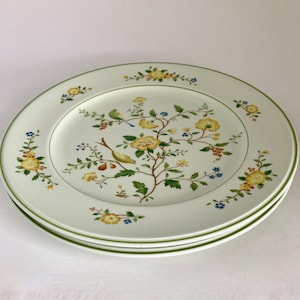 May include: Three white ceramic plates with a floral design. The plates feature a yellow and green floral pattern with birds and a green border. The plates are stacked on top of each other.