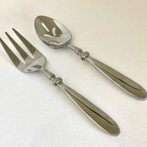 May include: A stainless steel salad fork and slotted spoon set. The fork has three tines, and the spoon has a slotted bowl. Both utensils have long, smooth handles with a decorative detail where the handle meets the utensil head.