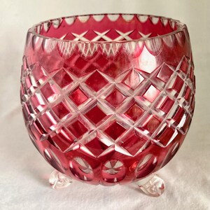 Puede incluir: Un cuenco de cristal tallado de color rojo rubí con un patrón de diamantes y un borde festoneado. El cuenco tiene tres pequeños pies transparentes. El cristal es transparente y probablemente se utiliza con fines decorativos.