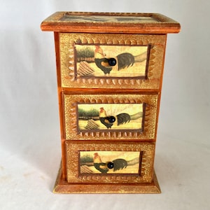 May include: A wooden chest of drawers featuring three stacked drawers, each adorned with a rooster illustration on a light background. The frame has a textured, golden-brown finish with scalloped edges around each drawer front and small black knobs.