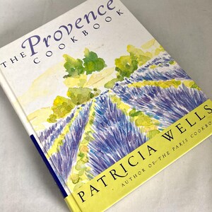 May include: A hardcover cookbook titled The Provence Cookbook by Patricia Wells. The cover features a watercolor illustration of purple lavender fields and green foliage against a white background, with a yellow band at the bottom.