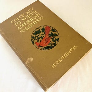May include: A vintage book titled "Color Key to North American Birds" by Frank M. Chapman. The cover is a muted gold color with gold lettering and a circular illustration of a red bird. The book is slightly worn.