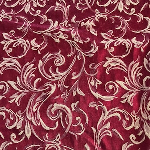 May include: A deep red velvet fabric featuring a repeating light beige damask pattern of swirling acanthus leaves and floral motifs. The texture of the fabric is soft and plush, with the intricate woven design creating a raised, tactile surface.