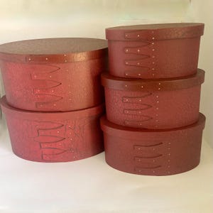 May include: A collection of six oval-shaped, red decorative boxes. The boxes are stacked, showcasing varying sizes. Each box features a textured surface and decorative stitching details, with a dark brown trim and lid.