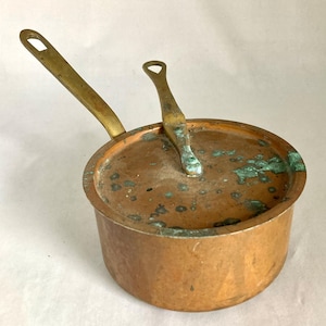 May include: A small copper saucepan with a lid and a long handle. The lid has a small handle. The copper has a patina with green and brown spots. The handles are brass-colored.