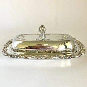 May include: A rectangular silver-plated butter dish with a removable lid. The base and lid feature an ornate embossed floral border. A decorative, sculpted finial sits atop the center of the lid, providing a handle for lifting the cover.