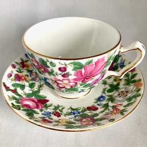 May include: A white teacup and saucer set with a floral pattern. The cup and saucer are decorated with pink, red, yellow, and blue flowers and green leaves. The rims are trimmed with gold.