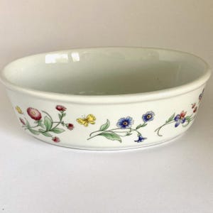 May include: A white ceramic oval bowl featuring a colorful botanical pattern around the exterior. The design includes various small flowers in shades of red, blue, and green, along with a single yellow butterfly depicted among the floral stems.