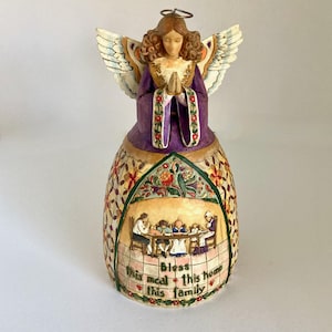 May include: A decorative angel figurine with brown hair and a silver halo, wearing a purple robe with floral trim. The skirt features a relief scene of a family dining at a table with the text, Bless this meal this home this family, in green.