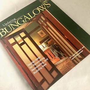 May include: A book titled "Updating America Bungalows" with a green cover and gold lettering. The cover features a photo of a room with a wooden door and bed. The book's subtitle reads "Design Ideas for Renovating, Remodeling, and Building New."