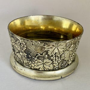 May include: Silver-toned wine coaster with a gold interior. The exterior features a detailed relief design of grape vines and leaves. The coaster has a circular base and a slightly flared rim, suitable for holding a wine bottle.