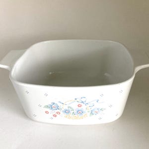 May include: A white square ceramic baking dish with integrated side handles. The exterior features a central floral basket motif in blue, green, and red, surrounded by a repeating pattern of small blue dots across the front surface.