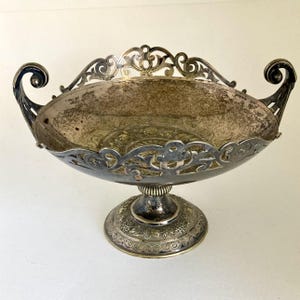 May include: An antique silver-toned centerpiece bowl with ornate scrollwork and a pedestal base. The bowl has a wide, oval shape with decorative handles and a detailed rim. The base features floral patterns.