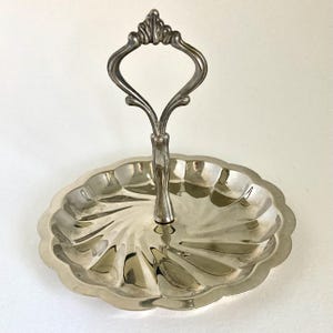 May include: Silver-toned, two-tiered serving tray with a scalloped edge and a decorative handle. The tray has a spiral design in the center and is made of a shiny metal. The handle is ornate and adds a touch of elegance to the piece.