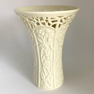 May include: A cream-colored ceramic vase featuring a flared, openwork lattice rim and a textured body decorated with embossed floral patterns. The cylindrical base transitions into a wide, decorative top with intricate cut-out detailing.