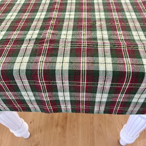 May include: A rectangular tablecloth featuring a classic plaid pattern in shades of forest green, deep red, and cream. The fabric displays a woven texture with thin gold metallic threads running vertically and horizontally throughout the design.