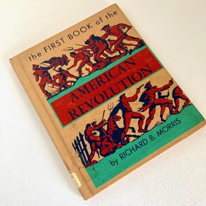 Może przedstawiać: Książka w twardej oprawie zatytułowana The First Book of the American Revolution autorstwa Richarda B. Morrisa. Beżowa okładka zawiera czerwone i zielone ilustracje żołnierzy w walce, w tym postacie z muszkietami i flagą na beżowym tle.