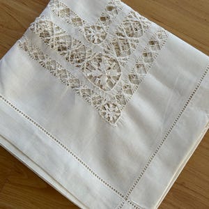 May include: A folded, off-white linen tablecloth with intricate lace detailing in the corner. The lace features a geometric pattern with floral accents. The edges of the tablecloth are finished with a delicate, stitched border.