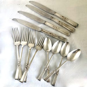 May include: A collection of vintage silverware, including knives, forks, and spoons. The silverware has a silver finish with decorative floral patterns on the handles. The knives are arranged at the top, with the forks and spoons below.
