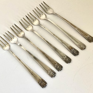 May include: A set of six silver-toned metal forks arranged diagonally. Each fork features a four-tine head and a slender handle that terminates in a decorative embossed floral pattern at the base. The metal surface shows a polished finish.