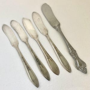 May include: A collection of silver butter knives. Five knives have a simple, curved blade design with decorative handles. One knife has a more ornate handle with floral detailing.