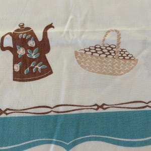 May include: A white fabric featuring a brown teapot with light blue and white floral accents, a tan woven basket filled with small round items, and a horizontal border design consisting of brown lines and a wide teal band at the bottom edge.