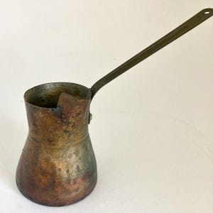 May include: A weathered copper coffee pot with a wide base, a narrow neck, and a long, flat metal handle attached with a single rivet. The exterior surface shows a dark, oxidized patina with visible color variations ranging from brown to green.