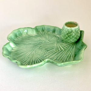 May include: A green ceramic serving dish and small cup. The dish is leaf-shaped with embossed pine needles and cones. The cup has a textured, pineapple-like design. The set is a light, glossy green.