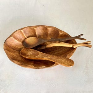 May include: A wooden serving bowl with a scalloped edge holds several wooden spoons of varying sizes and shapes. The bowl and spoons are made of a light brown wood.