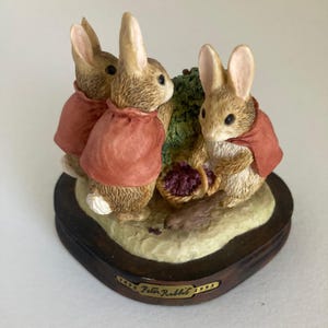 May include: A figurine featuring three brown rabbits wearing red jackets, gathered around a small basket of purple berries and a bird on a textured base. A gold plaque on the front of the dark wooden base reads 1893 Peter Rabbit 1993.