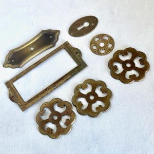 May include: An assortment of antique brass hardware components, including a rectangular frame, keyhole covers, decorative rosettes, and a rectangular plate. The pieces have a weathered, aged appearance, with a warm, golden-brown color.