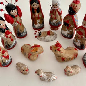 May include: A collection of hand-painted clay nativity figurines featuring human figures with black, white, or brown hair, some wearing red accents, alongside animal figures including sheep and cattle, and a small infant figure in a central manger.