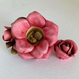 Może przedstawiać: Rzeźba różowej róży z pustym środkiem, w którym znajduje się miniaturowa figurka siedząca przy biurku komputerowym z klawiaturą i monitorem. Mniejszy, oddzielny pąk różowej róży znajduje się po prawej stronie głównego kwiatu.