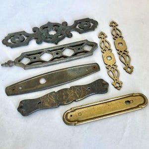 May include: A collection of antique metal hardware pieces in various shapes and finishes. Includes ornate, decorative pulls and plates in silver, brass, and bronze tones. Ideal for restoration projects or vintage decor.