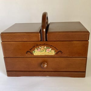 May include: A brown wooden sewing box with a central handle, two hinged top lids, and a single bottom drawer. The front features a floral decal with the text "Beautiful flowers will warm your heart" and the words "Floral House" in script.